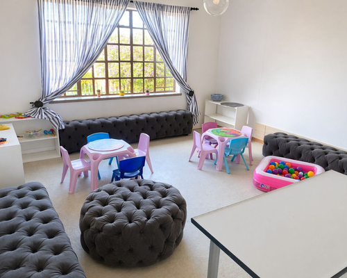 Playroom for kids with pink/blue plastic tables and chairs, a pink ball pit, tufted gray ottomans, white shelves, and a large window with striped blue curtains.