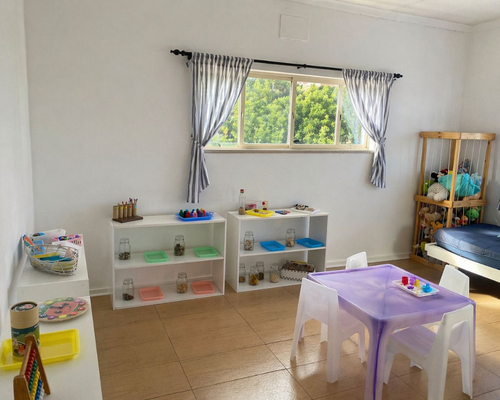 Playroom with white shelves of jars and toys, a purple table with white chairs, and a window with blue curtains.