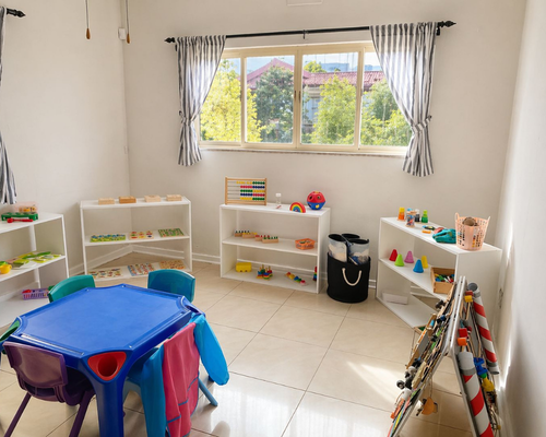 Bright children's playroom with white shelves of toys, a blue table and colored chairs, and a window with striped curtains.