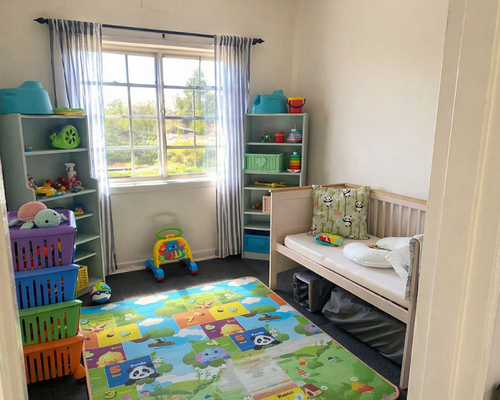 Bright children's playroom with a window, striped curtains, colorful toy shelves, a small bed, and a vibrant animal-pattern rug.