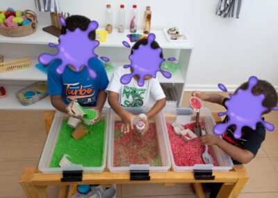Three kids at a wooden table play with bins of green and red beads, scooping and pouring in a sensory activity behind purple-covered faces.