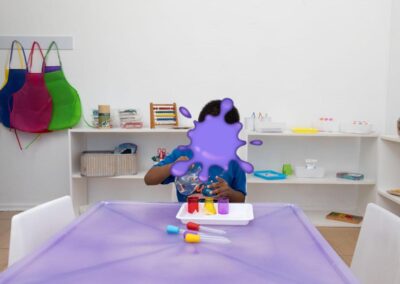 Child sits at a purple art table with small cups of colored paint, in a classroom with white shelves and colorful aprons on the wall behind them.