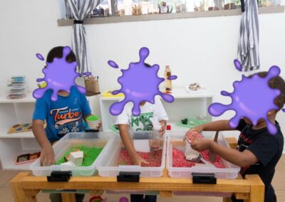 Three children stand at a wooden sensory table, scooping colorful beads into cups at a play station in a bright room