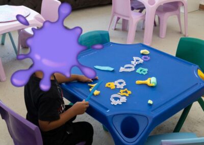 Child sits at a blue plastic table outdoors, using a blue sculpting tool with assorted cookie cutters and tools on the table; face covered by a purple splash image mask.