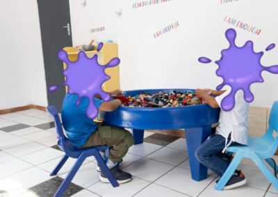 Two children sit at a blue round Lego table, sorting and playing with colorful bricks in a bright playroom.