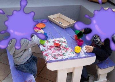 Two children sit at a purple-painted table, painting with colorful cups and a floral cloth laid on the table (faces obscured). Includes a wooden tray and paint splatters around.