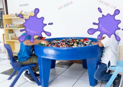 Two children building with LEGO bricks at a large blue round table in a bright playroom.