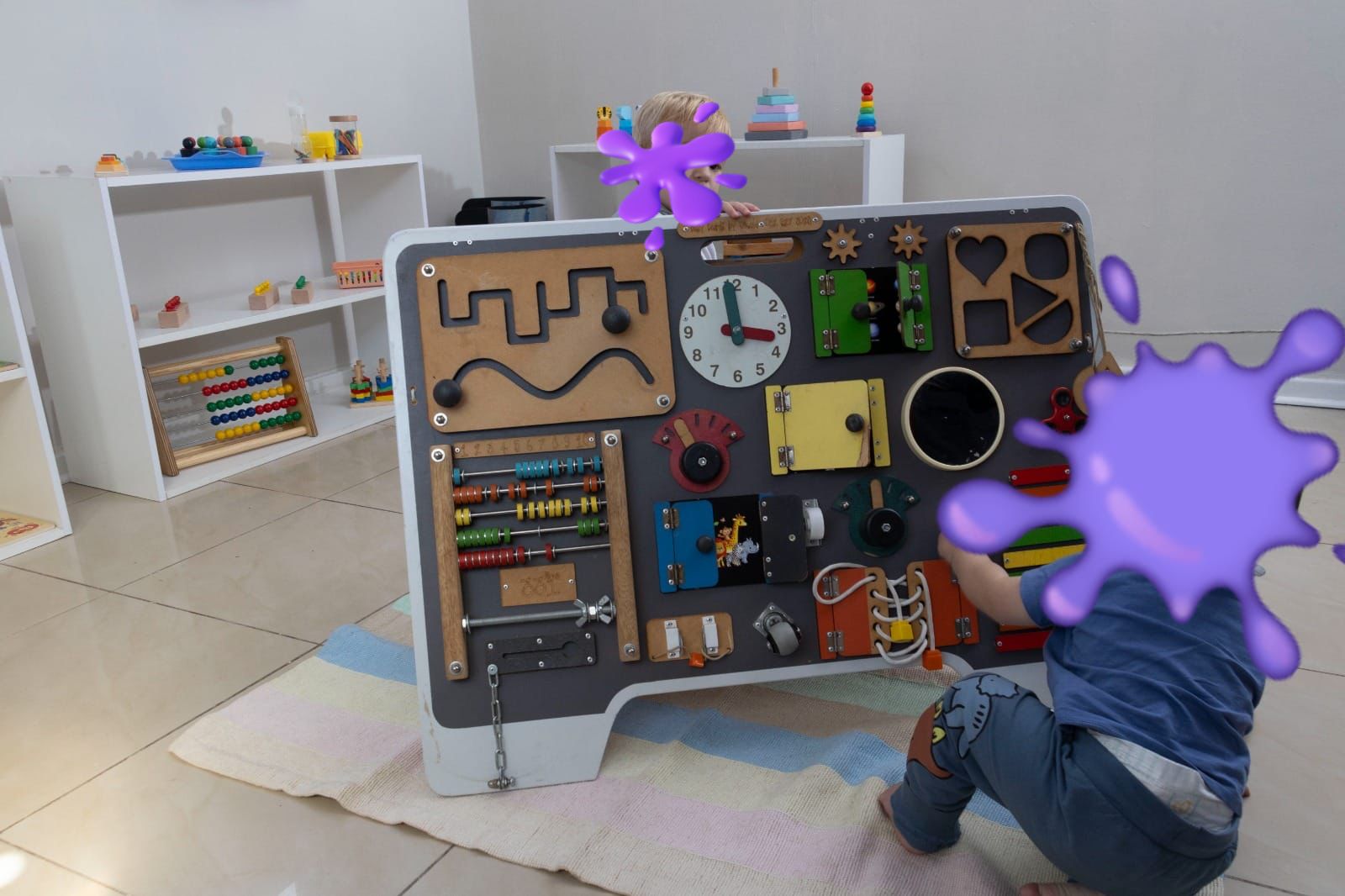 Toddler kneels at a large multi‑sensory activity panel with clocks, gears, beads, and levers in a bright playroom.