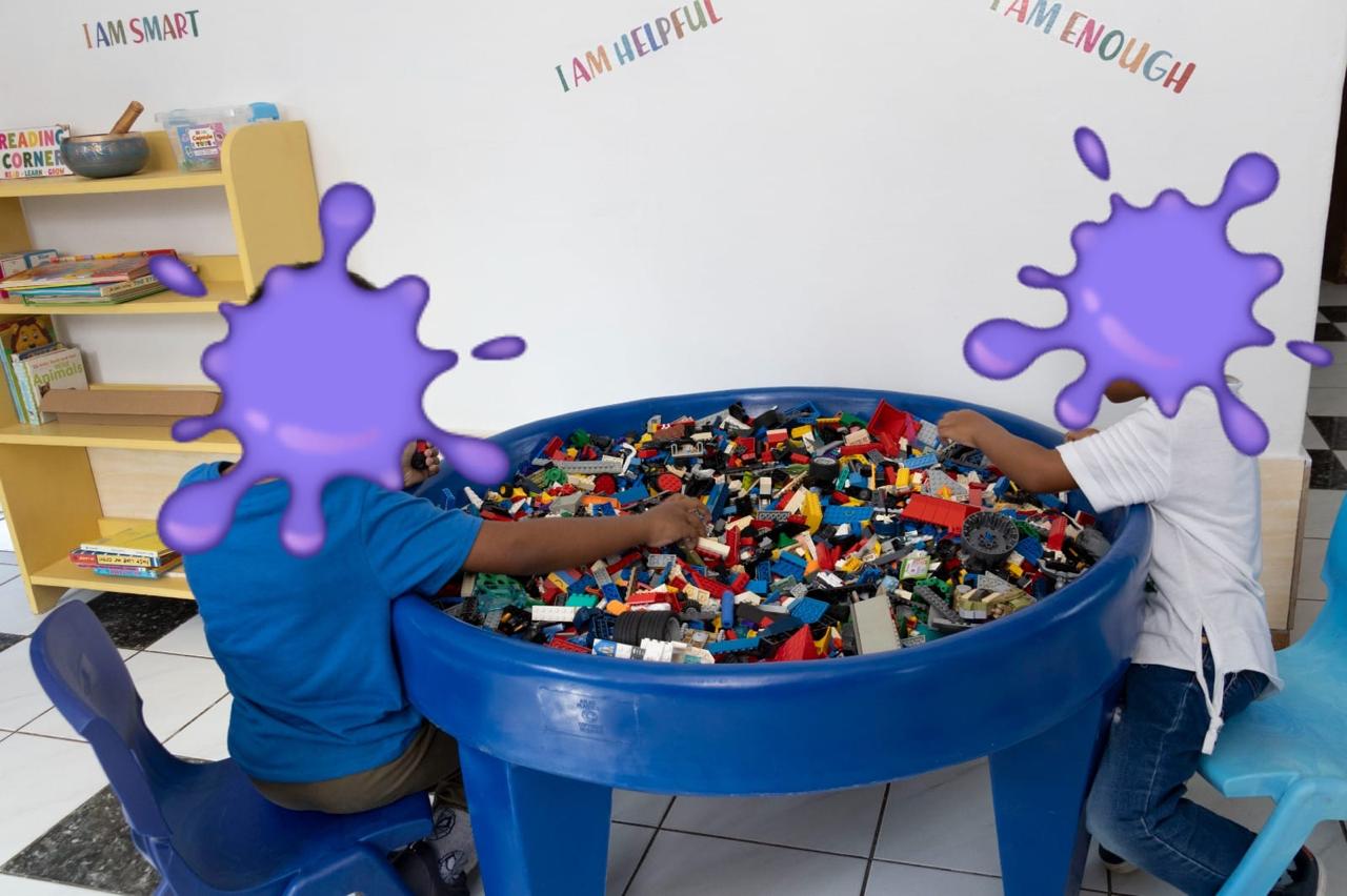 Two children sit on blue chairs at a round blue tub filled with colorful LEGO bricks, reaching in to build in a classroom setting.