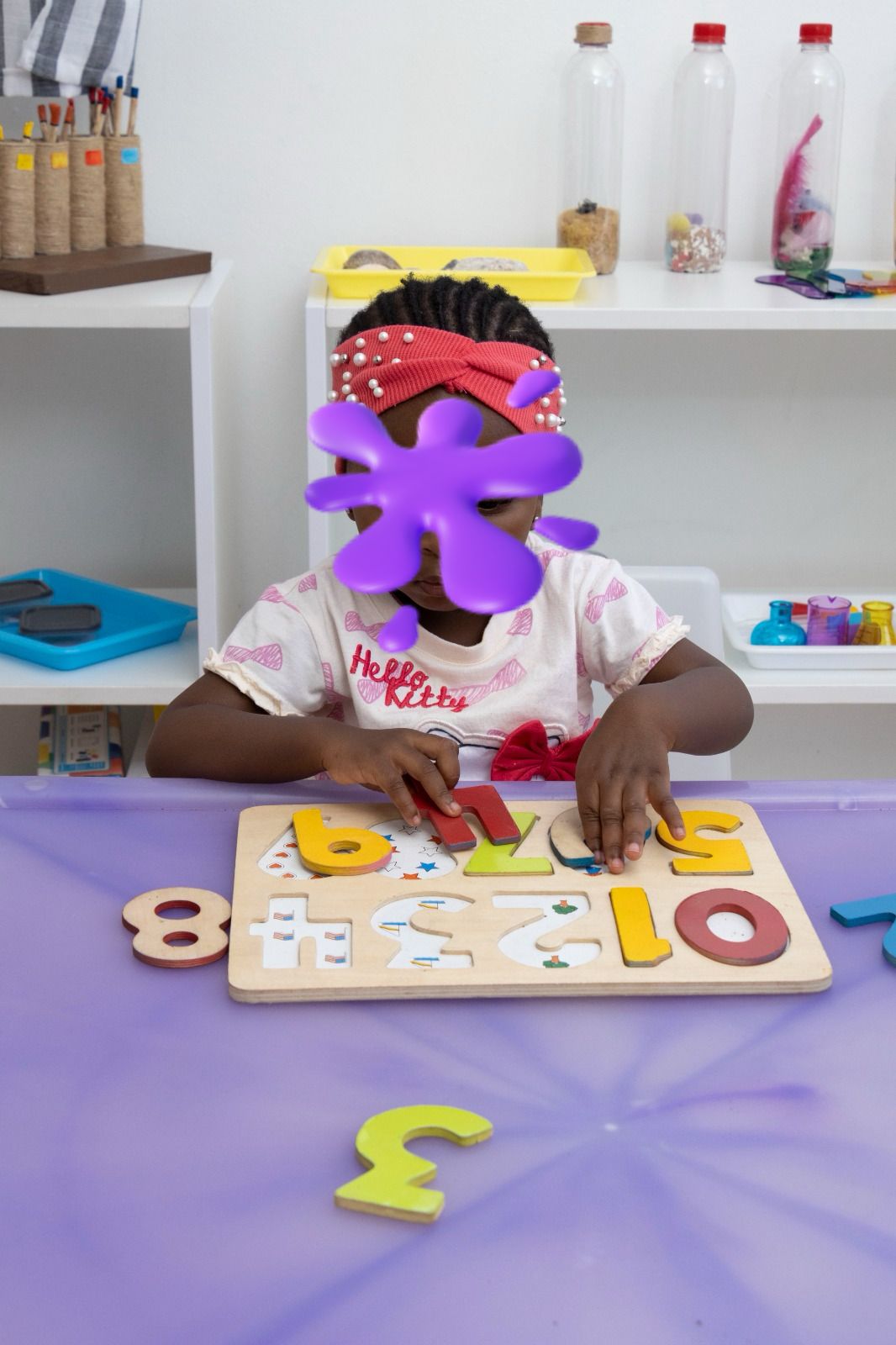 Young child sits at a purple table, working with a wooden number/letter puzzle on a board in a bright classroom.