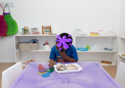 Child at a crafts table in a bright classroom, wearing a purple star-shaped mask while arranging colorful shapes on a tray of rocks and paper pieces in front of them at a purple table.
