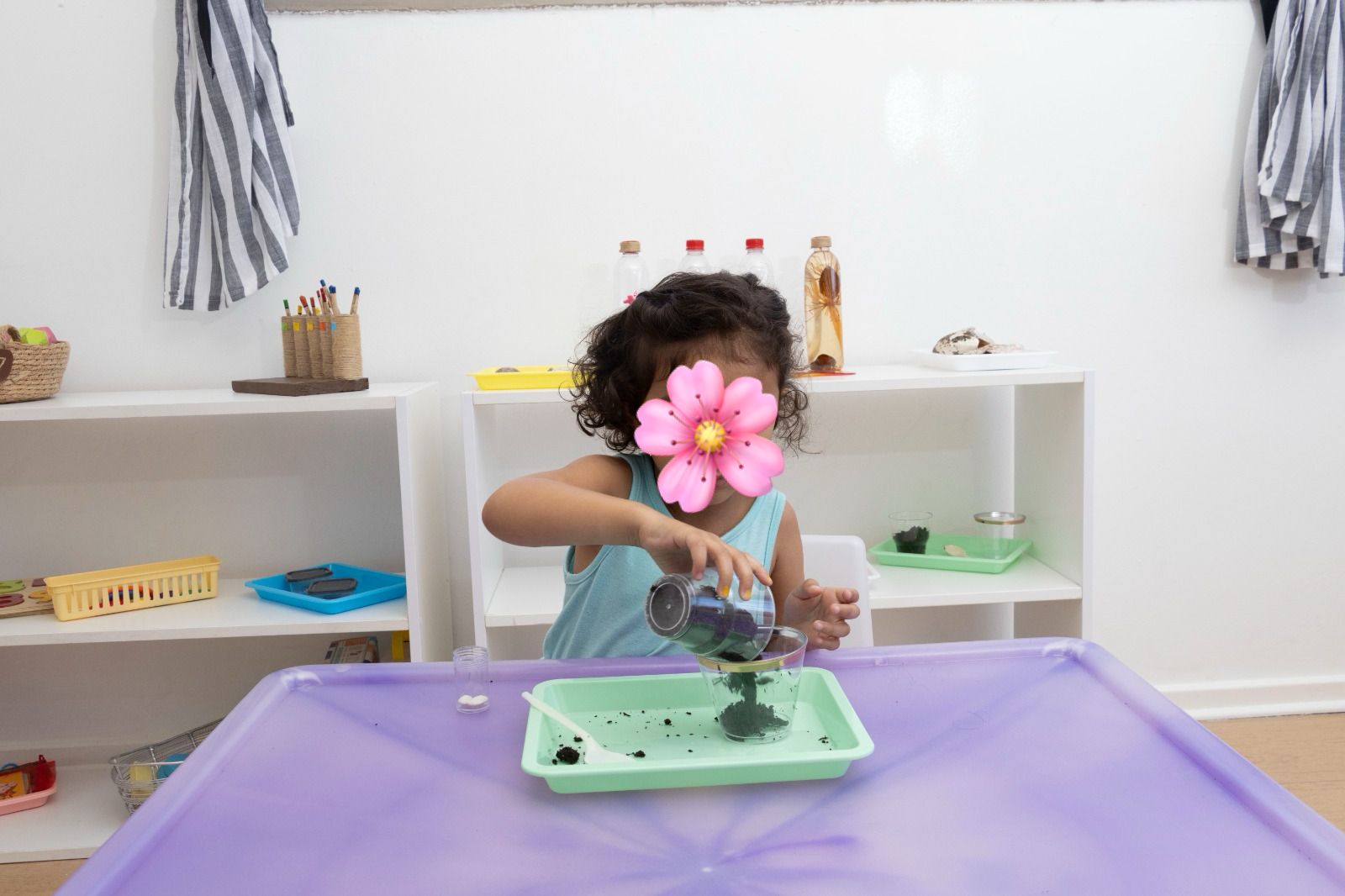 Child at a sensory play station pours dark liquid from a cup into a green tray on a purple table, a pink flower obscuring her face.