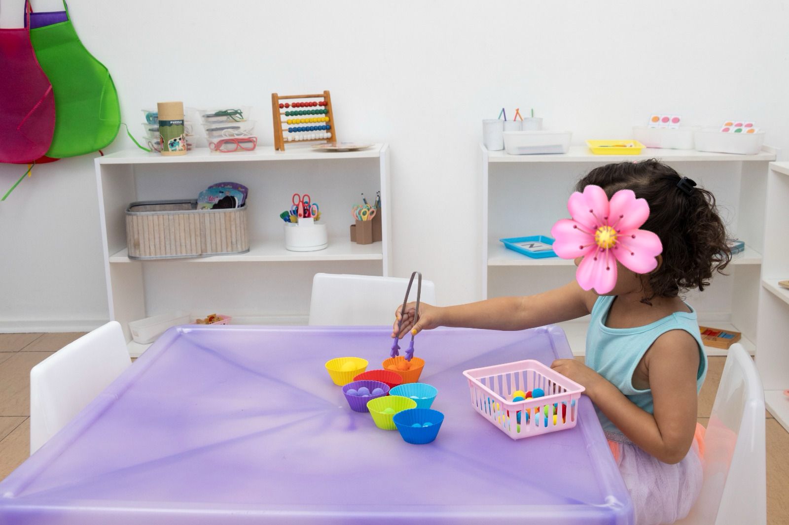 Young girl in a blue top sits at a purple craft table, using tongs to pick colorful cupcake liners from a set of bowls and a pink basket of beads in a bright classroom.