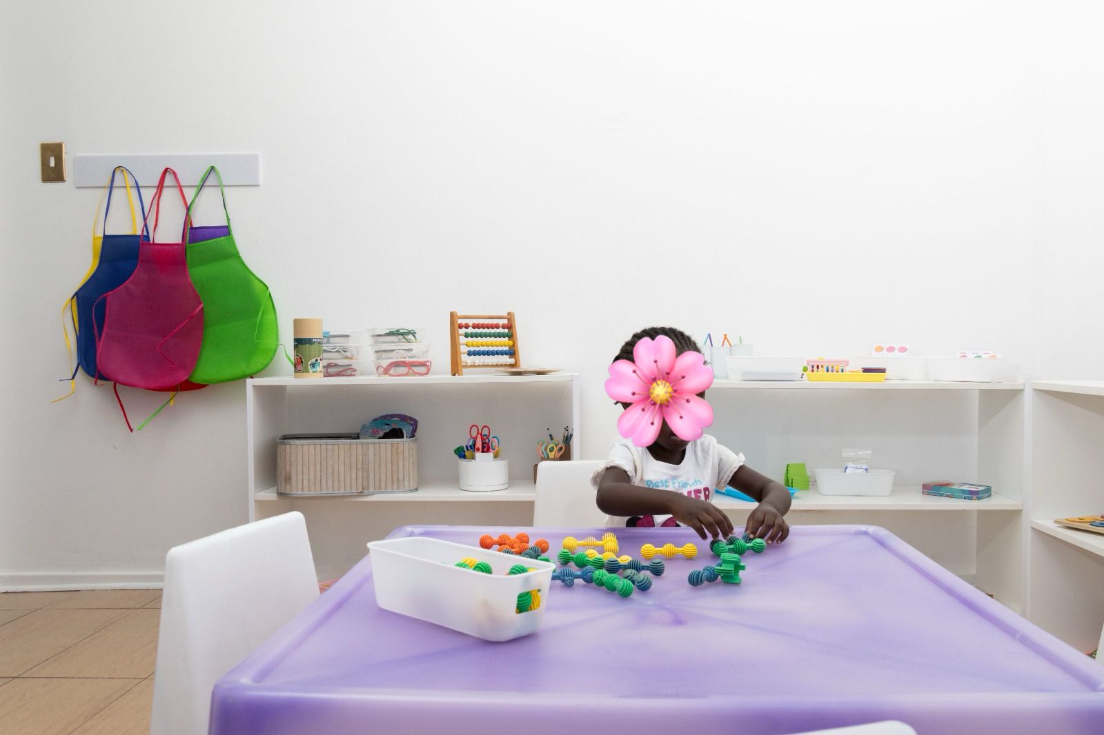 Child sitting at a purple activity table sorting colorful beads in a bright classroom; a flower covers their face for privacy.