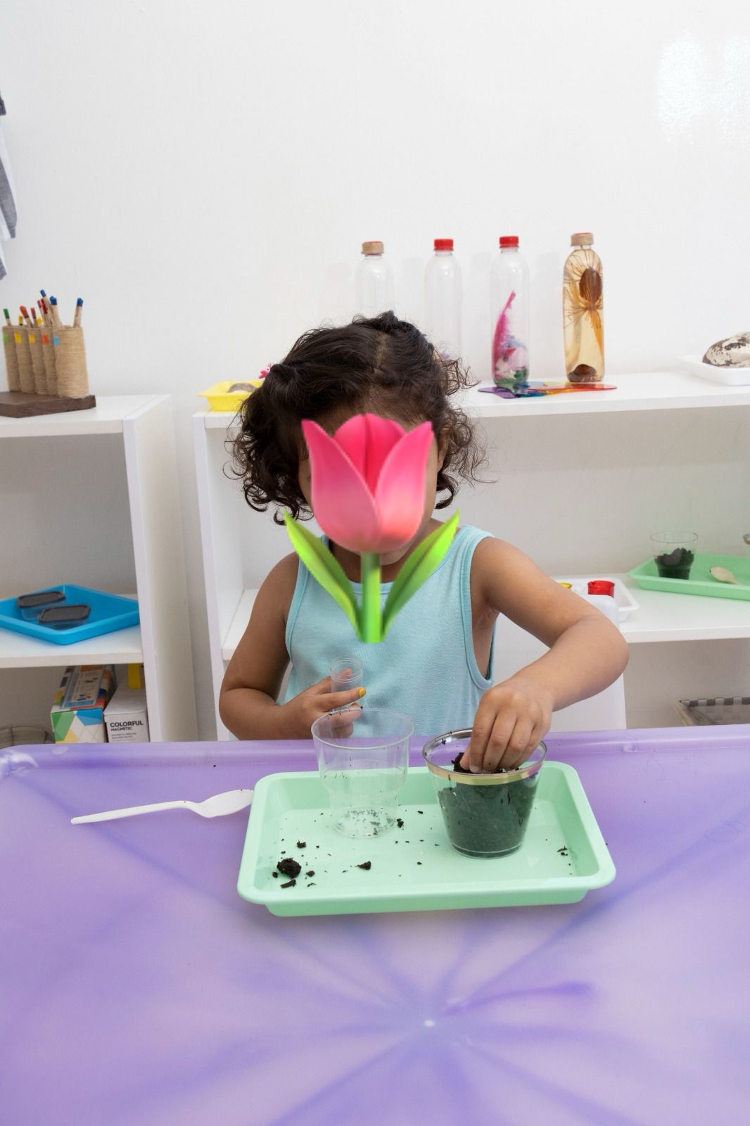 Child at a craft table holds a pink tulip in front of their face while working with soil in a green cup on a purple table.