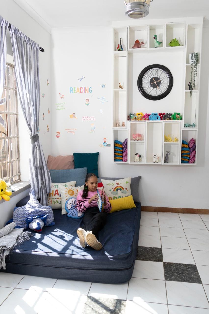 Girl reading a book on a dark blue mattress in a bright room with white wall shelves, a large clock, and colorful cushions.