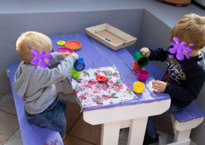 Two young children sit at a purple-painted wooden table, pouring colorful paint from cups onto a napkin-covered surface, crowded with small bowls and a wooden tray nearby (faces obscured).