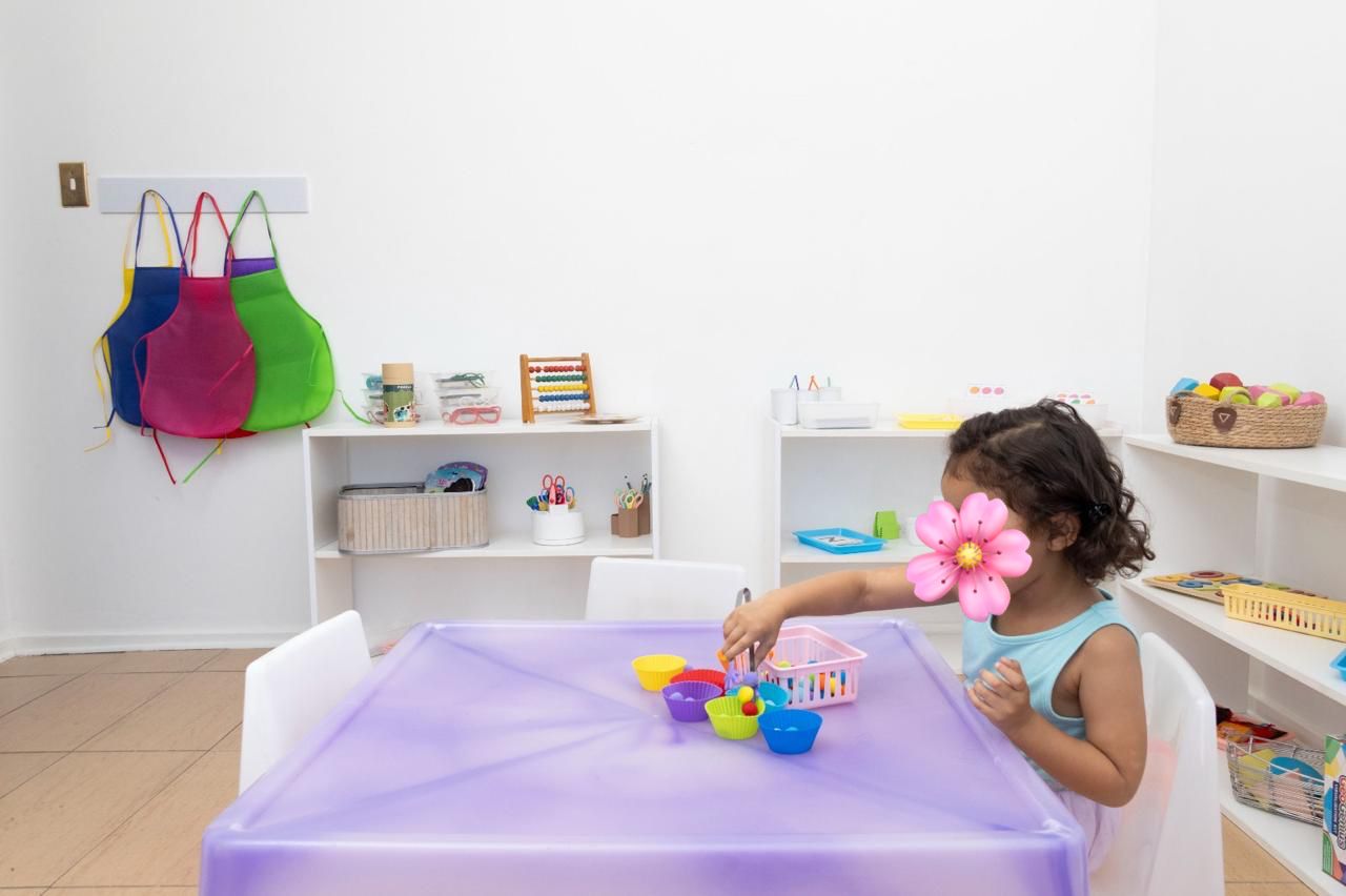 Child sits at a lavender table, playing with colorful plastic bowls and spoons in a bright, organized playroom.