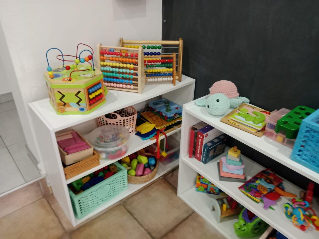 WhatsApp Image 2026-08-17 at 235031 - Margies Montessori Inspired Roodepoort Colorful children's toys on white shelves, including bead mazes, a wooden abacus, baskets of blocks, and books on display.