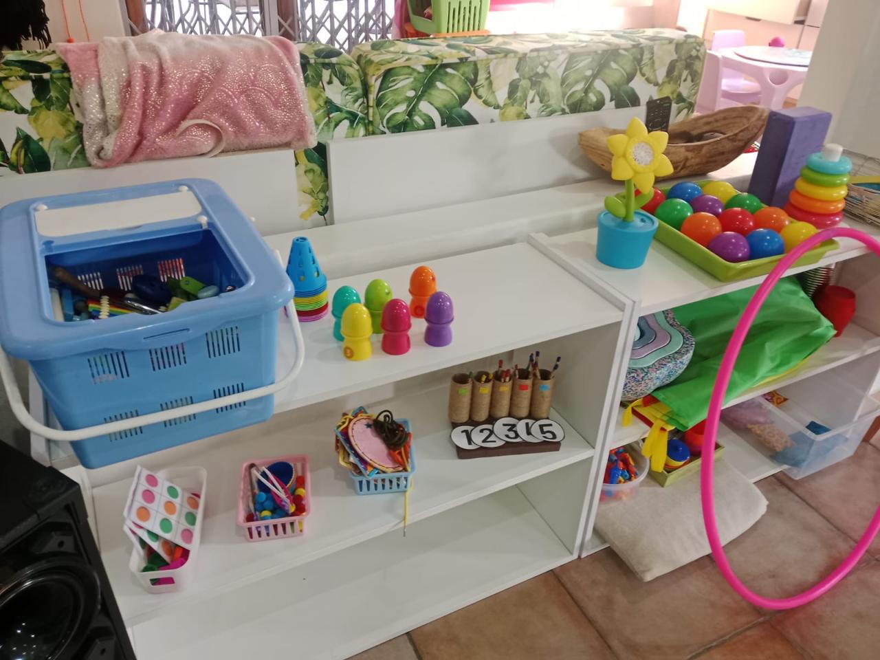 WhatsApp Image 2026-08-17 at 235032 1 - Margies Montessori Inspired Roodepoort Children's play area with white shelving, a blue plastic basket of toy tools, and colorful stacking toys on display.