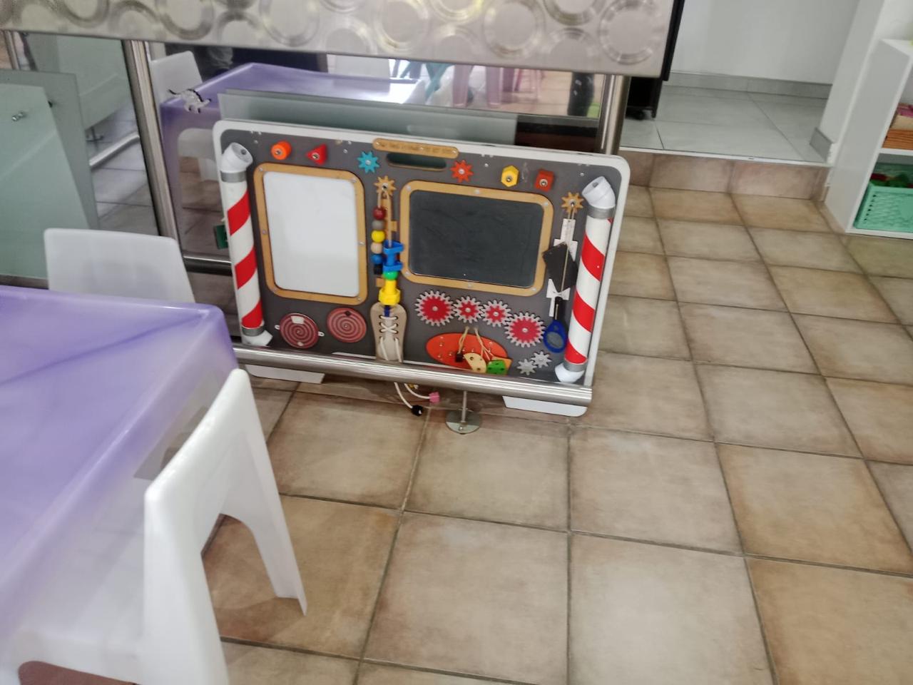 WhatsApp Image 2026-08-17 at 235032 - Margies Montessori Inspired Roodepoort Freestanding children's activity board with gears, beads, and sliders in a tiled room near a purple table and white chair