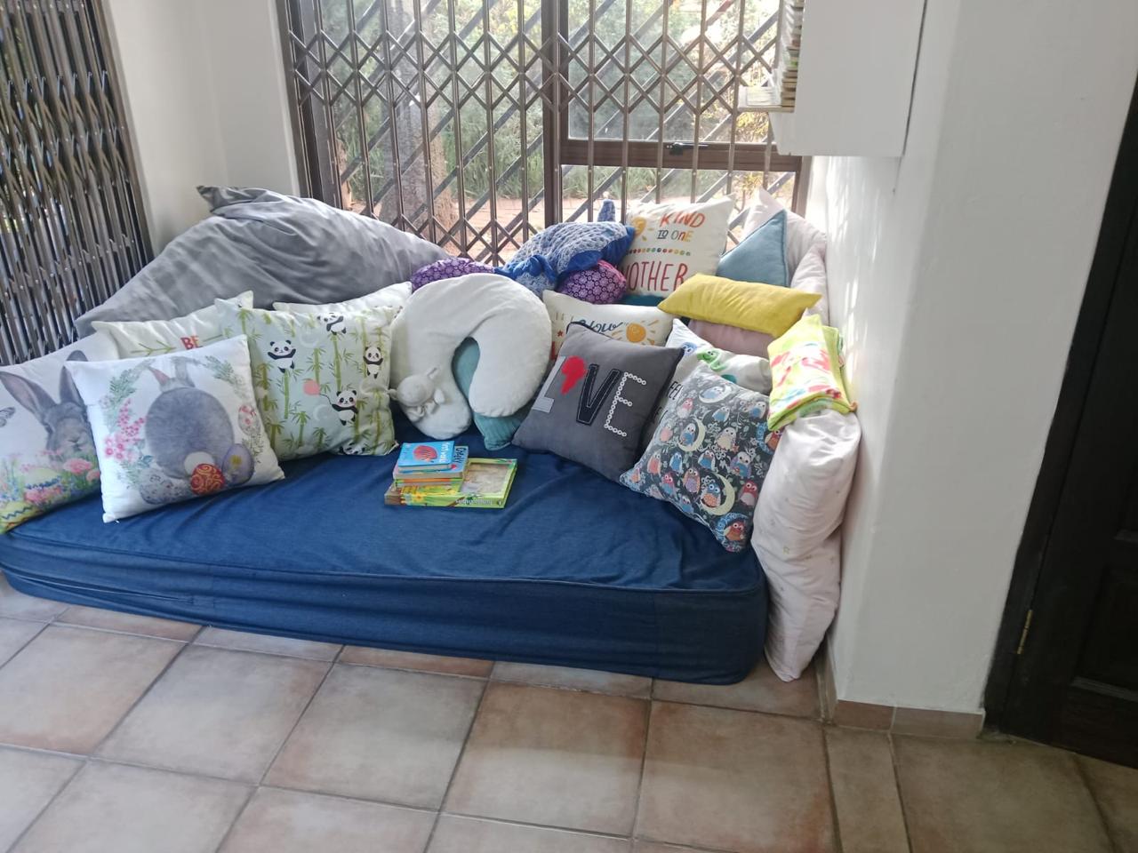 WhatsApp Image 2026-08-17 at 235033 2 - Margies Montessori Inspired Roodepoort Cozy blue cushioned bench on a balcony with colorful decorative pillows and a few books on the seat edge near a metal railing barrier.