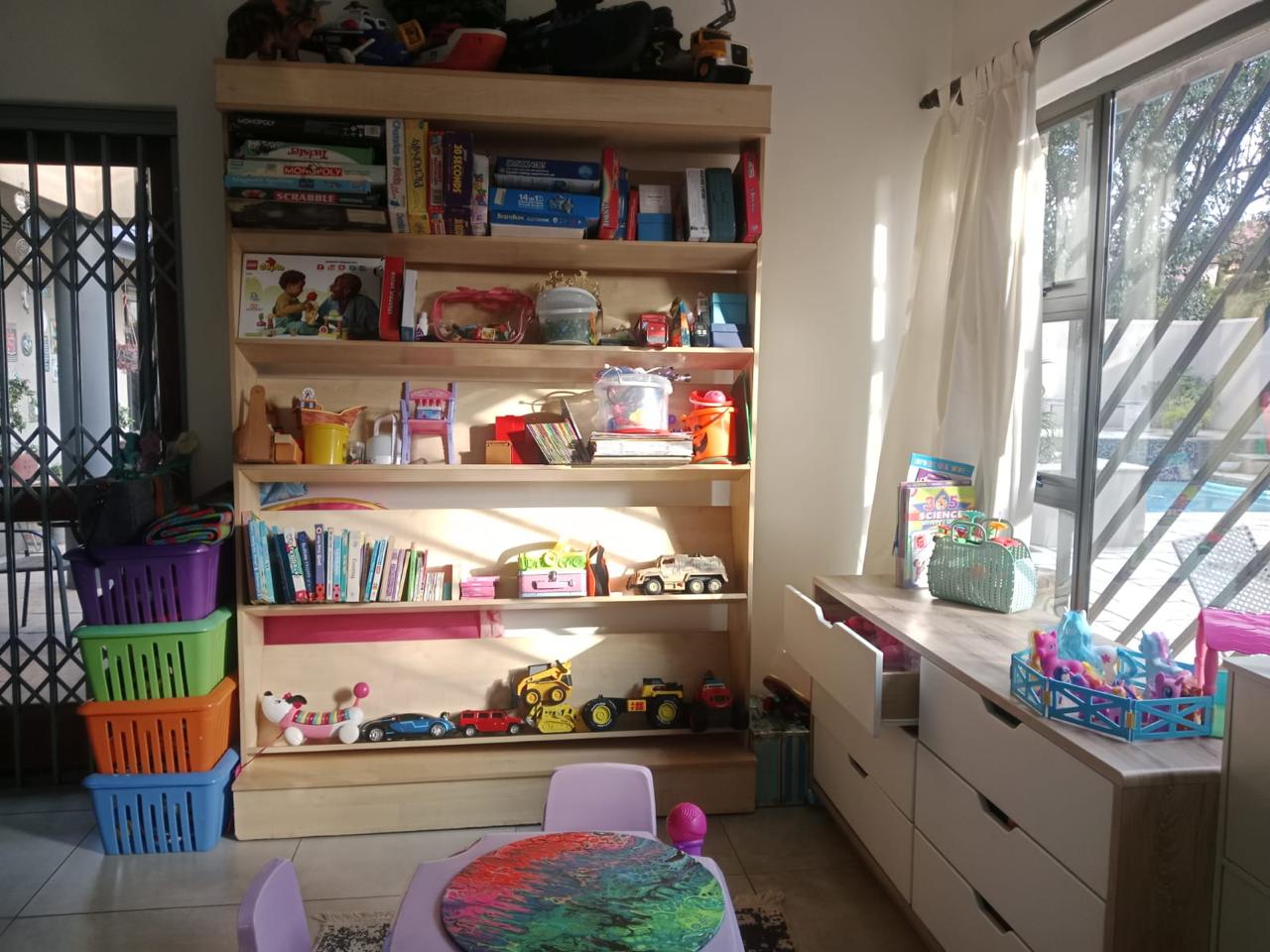WhatsApp Image 2026-08-17 at 235034 - Margies Montessori Inspired Roodepoort Wooden bookshelf laden with toys, books, and games in a bright playroom; colorful bins stack on the left and a window with curtains on the right.